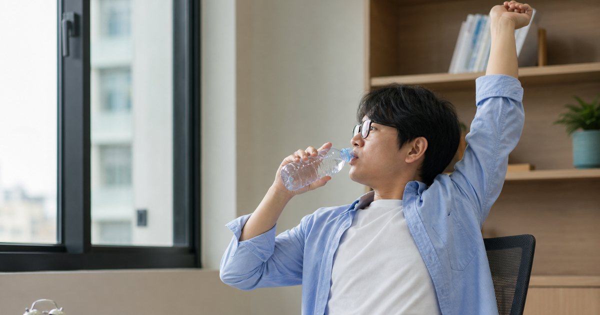 A tutor stretching and drinking water beside a window in a quiet study room, a stack of workbooks and a small analog clock on the desk, midday light